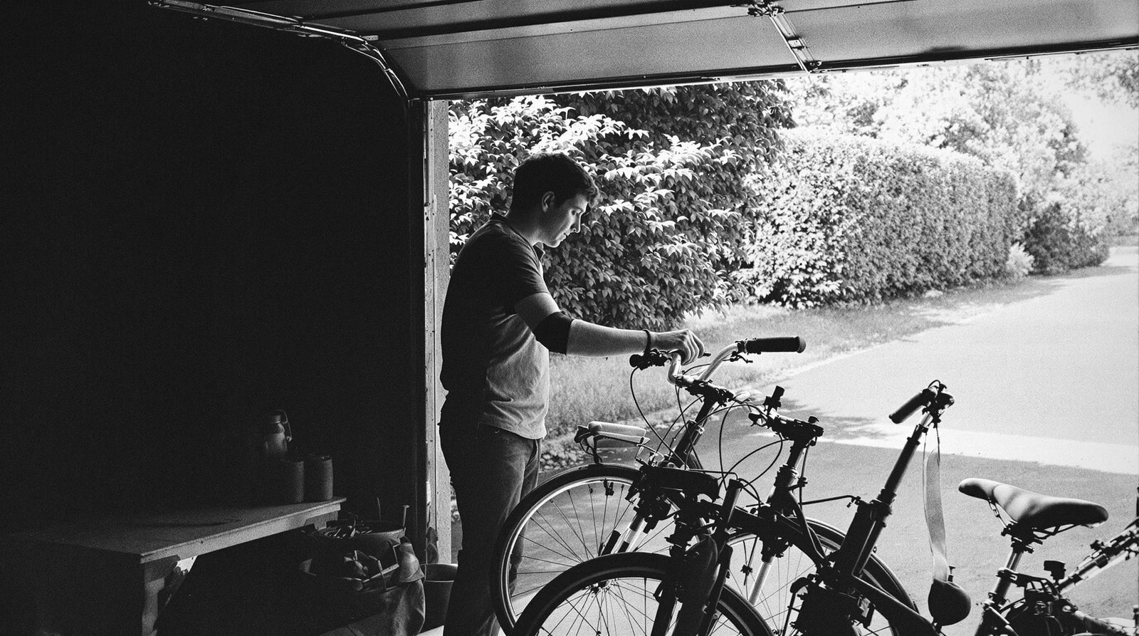 Person im Halbprofil an einem offenen Garagentor, arbeitet an einem Fahrrad, dahinter eine sonnige Einfahrt
