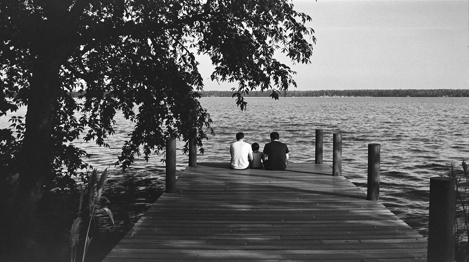 Eine Familie sitzt am Ende eines Holzstegs am See, von hinten gesehen