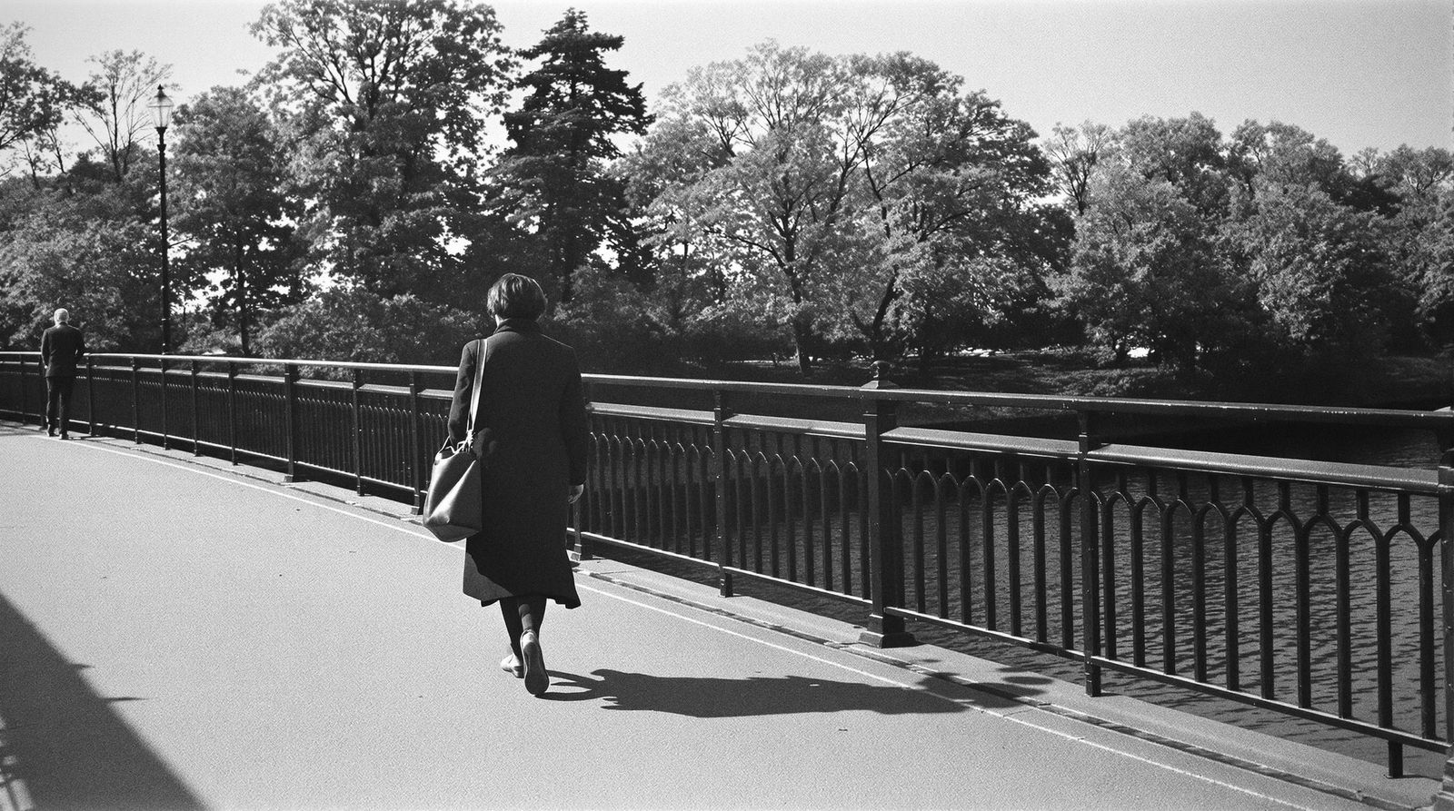 Eine Frau mit Umhängetasche geht über eine Fußgängerbrücke am Fluss, von hinten gesehen